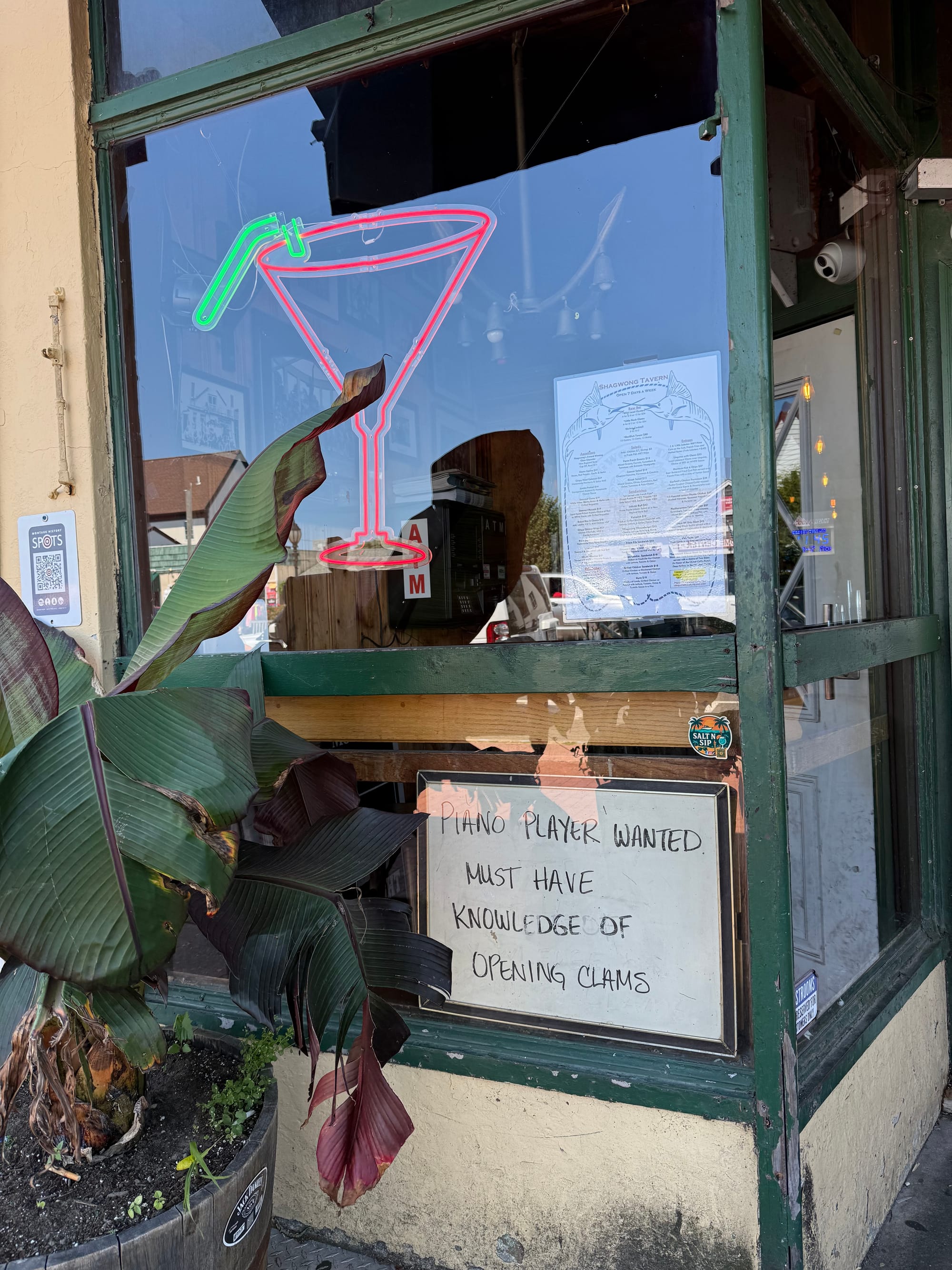 A photo depicting a neon martini glass in a bar window with a want ad. Posted in the window of a cocktail lounge and seafood restaurant. Text in caption.